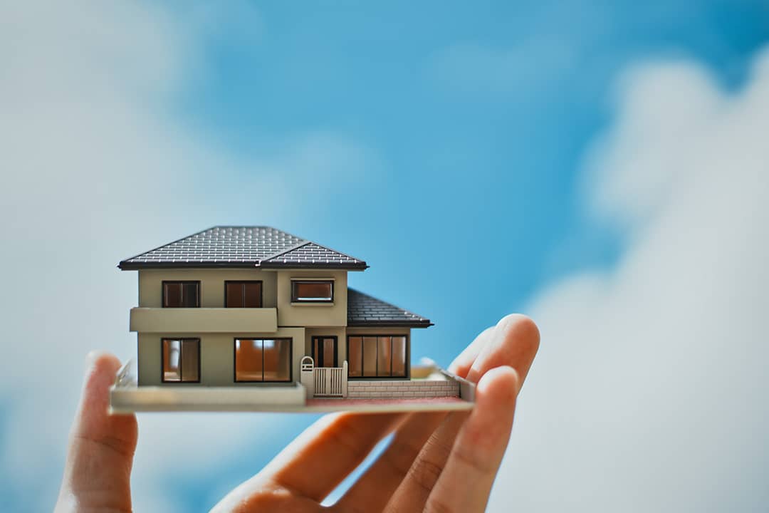 A person's hand holding a miniature house with the bright sky in the background.